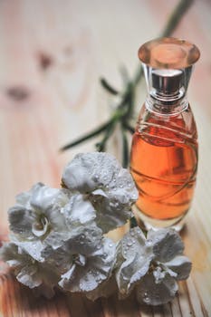 Glass perfume bottle alongside dew-kissed white carnation flowers on wood surface.