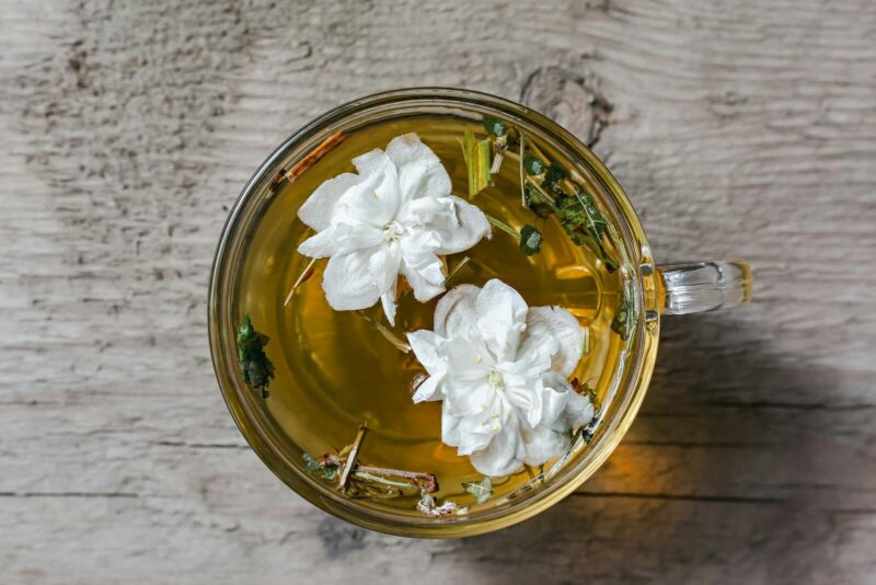 From above of aromatic herbal tea with fresh Arabian jasmine flowers in glass cup placed on wooden table in daylight